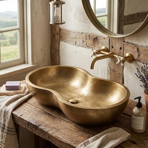 May include: A gold-toned, hammered metal vessel sink with an irregular, kidney-bean shape. The sink features a central drain and is mounted on a rustic wooden surface, paired with a wall-mounted brass faucet with two white cross-handle knobs.