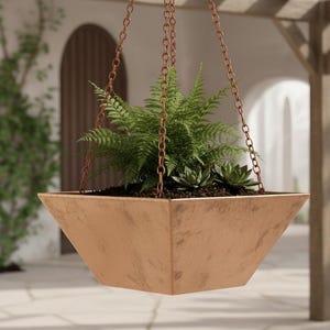 Op de afbeelding: Een vierkante, koperkleurige hangende plantenbak gevuld met groene planten. De plantenbak hangt aan kettingen en hangt voor een witte stucwerkmuur met boogvensters en een houten pergola.