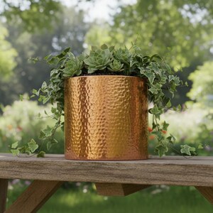 Pode incluir: Um vaso de metal martelado cor de cobre cheio de suculentas verdes e hera pendurada. O vaso cilíndrico está sobre uma mesa de madeira, com um fundo de jardim desfocado. O vaso adiciona um toque de elegância a qualquer espaço exterior.