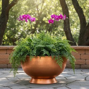 Puede incluir: Una gran maceta de cobre martillado llena de helechos verdes y vibrantes orquídeas moradas. La maceta se encuentra en un patio de piedra, con una pared de ladrillo y árboles en el fondo. Un acento decorativo para exteriores.
