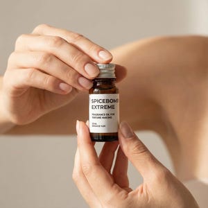 May include: A small, brown glass bottle with a silver cap, labeled "SPICEBOMB EXTREME" and "FRAGRANCE OIL FOR PERFUME MAKING." The bottle contains 10 ml of fragrance oil. The bottle is held between two hands.