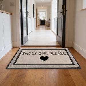 Pode incluir: Tapete retangular com o texto "SHOES OFF, PLEASE" e um coração preto. O tapete tem um centro bege com uma borda preta, colocado em um piso de madeira em um corredor.