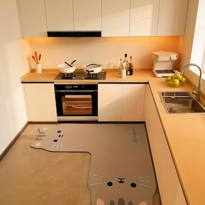 Puede incluir: Una cocina moderna con armarios blancos, un fregadero de acero inoxidable y un horno empotrado. Una alfombra gris con forma de gato está en primer plano. La alfombra tiene las palabras "HAPPY CAT" impresas. Dos ollas blancas están en la estufa.