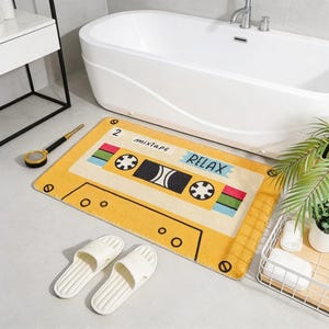 Op de afbeelding: Gele badmat in de vorm van een cassettebandje met de woorden "RELAX" en "mixtape" erop gedrukt. De mat heeft een retro-ontwerp met zwarte, rode, groene en blauwe accenten. Witte slippers liggen in de buurt van de mat, wat wijst op een badkamer.