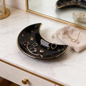 K&ouml;nnte beinhalten: Schwarze, halbmondf&ouml;rmige Schmuckschale mit goldenen Verzierungen und Sternen. Auf der Schale sind mehrere Ringe und ein Armband ausgestellt. Ein kleiner beiger Beutel und eine Halskette liegen neben der Schale auf einer wei&szlig;en Oberfl&auml;che.