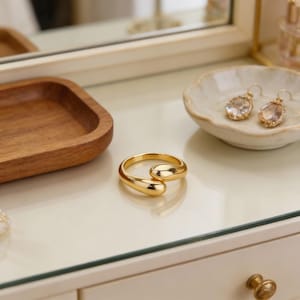 Op de afbeelding: Een goudkleurige ring met een uniek, vloeiend ontwerp. De ring ligt op een wit oppervlak, naast een kleine houten tray en een decoratief schaaltje met een paar oorbellen. De oorbellen hebben een heldere, druppelvormige steen.