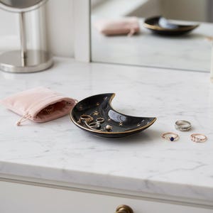 K&ouml;nnte beinhalten: Eine schwarze, halbmondf&ouml;rmige Schmuckschale mit goldenen R&auml;ndern und Sternen. Mehrere Ringe sind auf der Schale ausgestellt, neben einem rosa Samtbeutel. Die Schale steht auf einer wei&szlig;en Marmoroberfl&auml;che.