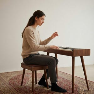 Puede incluir: Un piano digital de madera marrón oscuro con un banco a juego. El piano tiene un diseño moderno con patas anguladas. Las teclas son blancas y negras. El banco tiene un asiento de cuero marrón. El piano está sobre una alfombra estampada. Mide aprox. 120 cm de largo.