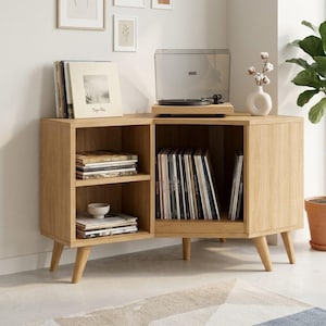 Puede incluir: Consola de tocadiscos de madera clara con un tocadiscos en la parte superior y espacio para guardar discos de vinilo. La consola tiene un diseño moderno de mediados de siglo con patas anguladas y estanterías abiertas. Un pequeño jarrón blanco con tallos decorativos está cerca.