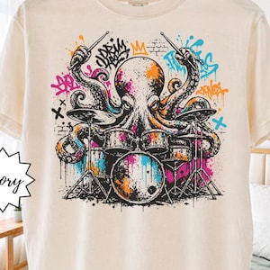 May include: Ivory-colored t-shirt featuring a colorful octopus playing a drum set. The octopus is depicted with orange, purple, and blue accents, with graffiti-style art in the background. The shirt is labeled "Super Soft."