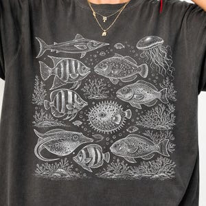 以下が含まれることがあります： 様々な魚と海洋生物の白いイラストが描かれた濃いグレーのTシャツ。サメ、クラゲ、様々な種類の魚、サンゴ、海藻が含まれています。「Pepper」の文字が左下に印刷されています。