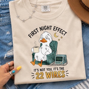 Könnte beinhalten: Ein cremefarbenes T-Shirt mit einer Grafik einer weißen Ente, die in einem blaugrünen Sessel sitzt, an medizinische Kabel und einen Monitor angeschlossen ist und eine Kaffeetasse hält. Der Text lautet: FIRST NIGHT EFFECT IT'S NOT YOU, IT'S THE 22 WIRES.