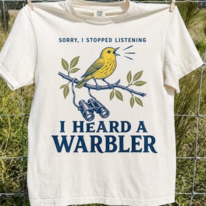 May include: An ivory t-shirt featuring a graphic of a yellow warbler perched on a branch with binoculars hanging below. The text reads, "SORRY, I STOPPED LISTENING I HEARD A WARBLER" in dark blue lettering, centred on the front of the garment.