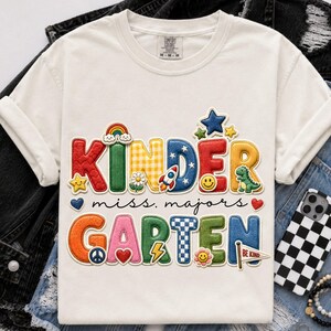 May include: An ivory t-shirt featuring a colourful graphic with the text "KINDER miss. majors GARTEN". The design includes various icons like a rainbow, stars, a rocket, a dinosaur, a peace sign, a heart, and a smiley face in multiple colours.