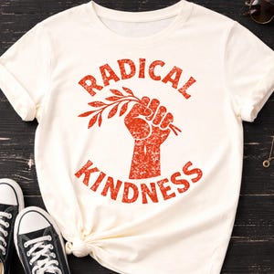 May include: Ivory-colored t-shirt with the words "RADICAL KINDNESS" in red, featuring a red fist holding a sprig. The shirt is paired with black sneakers with white laces. Sunglasses and a necklace are also visible.