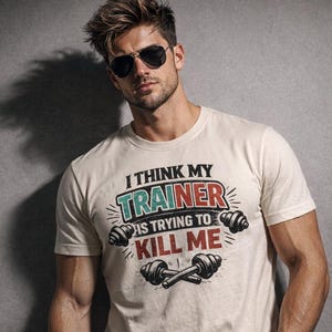 Op de afbeelding: Een crèmekleurig T-shirt met korte mouwen met de tekst I THINK MY TRAINER IS TRYING TO KILL ME in zwarte, rode en blauwgroene letters, vergezeld van grafische illustraties van twee halters die horizontaal en gekruist onder de tekst staan.