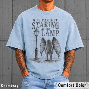 May include: A light blue short-sleeve t-shirt featuring a graphic print of a winged creature standing next to a street lamp. The text reads "GOT CAUGHT STARING AT THE LAMP". The creature has prominent red eyes and large, dark, textured wings.