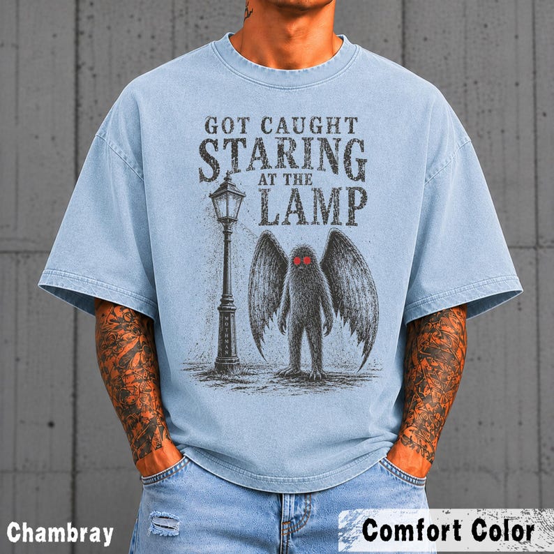 May include: A light blue short-sleeve t-shirt featuring a graphic print of a winged creature standing next to a street lamp. The text reads "GOT CAUGHT STARING AT THE LAMP". The creature has prominent red eyes and large, dark, textured wings.