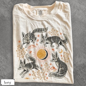 Puede incluir: Una camiseta color marfil con una ilustración en tinta negra de tres zorros en varias poses rodeando un gráfico central de sol y luna dorados. El diseño incluye pequeñas estrellas naranjas, insectos y ramitas botánicas.