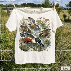 May include: An ivory short-sleeve t-shirt featuring a detailed graphic print of multiple ducks swimming in a pond. The illustration includes aquatic plants, lily pads, dragonflies, and small frogs, rendered in natural tones of green, brown, and blue.