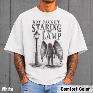 May include: A white short-sleeve t-shirt featuring a black graphic illustration of a winged creature standing next to a street lamp. The text above the graphic reads "GOT CAUGHT STARING AT THE LAMP" in a distressed black font. The creature has red eyes.