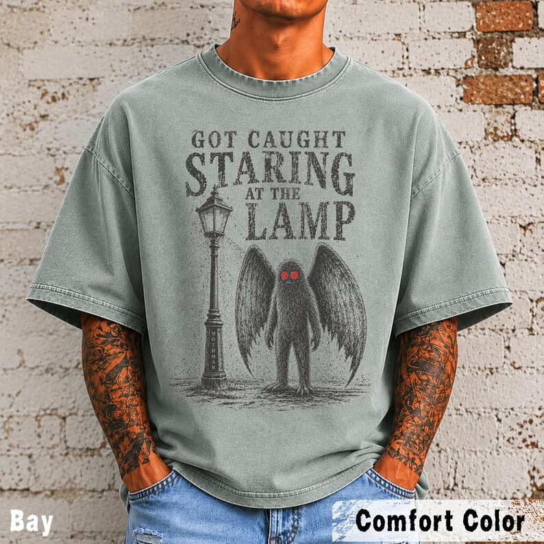May include: A sage green short-sleeve t-shirt featuring a graphic print of a winged creature standing next to a street lamp. The text reads "GOT CAUGHT STARING AT THE LAMP". The creature has dark fur and bright red eyes, and the design is in black ink.