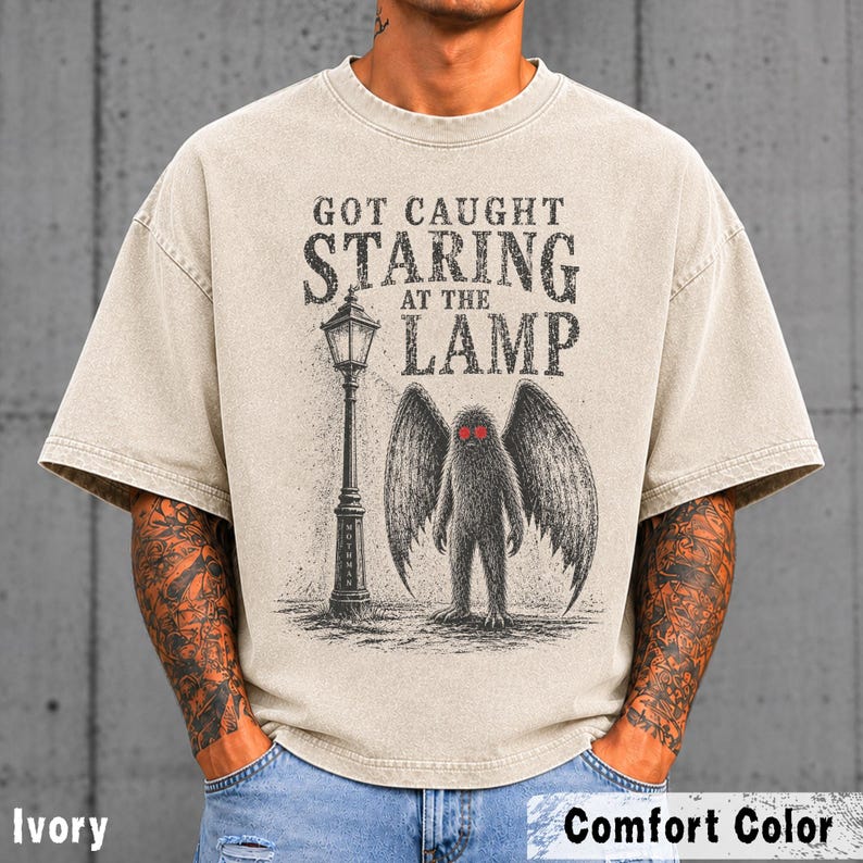 May include: An ivory short-sleeve t-shirt featuring a black graphic print of a winged creature standing next to a street lamp. The text reads GOT CAUGHT STARING AT THE LAMP. The creature has prominent red eyes and large, detailed feathered wings.