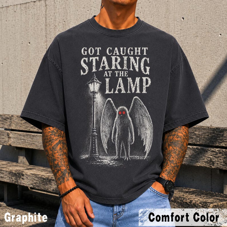 May include: A charcoal gray short-sleeve t-shirt featuring a graphic print of a winged creature standing next to a street lamp. The text reads GOT CAUGHT STARING AT THE LAMP. The creature has large wings and two bright red eyes in a sketch style.