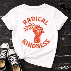 May include: White t-shirt with the words "RADICAL KINDNESS" in red, featuring a fist holding a branch. The shirt is paired with black sneakers with white laces. Sunglasses and a necklace are also visible.