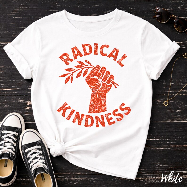 May include: White t-shirt with the words "RADICAL KINDNESS" in red, featuring a fist holding a branch. The shirt is paired with black sneakers with white laces. Sunglasses and a necklace are also visible.