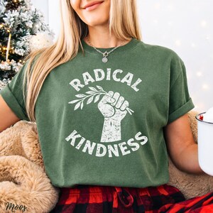 May include: Green t-shirt with the words "RADICAL KINDNESS" in white, along with a graphic of a clenched fist holding a sprig. The shirt has rolled-up sleeves and a crew neck. The model is wearing a silver necklace and red and black plaid pants.
