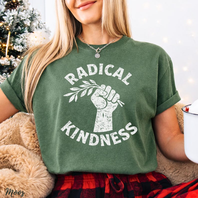 May include: Green t-shirt with the words "RADICAL KINDNESS" in white, along with a graphic of a clenched fist holding a sprig. The shirt has rolled-up sleeves and a crew neck. The model is wearing a silver necklace and red and black plaid pants.