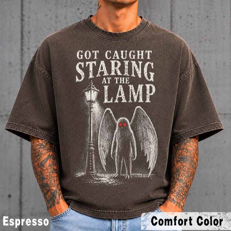 May include: An espresso-colored short-sleeve t-shirt featuring a graphic print of a winged creature with red eyes standing next to a street lamp. The text reads GOT CAUGHT STARING AT THE LAMP. The design is rendered in white and red ink on the dark fabric.