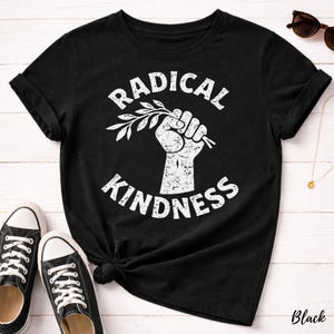 May include: Black t-shirt with the words "RADICAL KINDNESS" in a white, distressed font. A white fist holds a sprig of leaves. The shirt is paired with black sneakers.