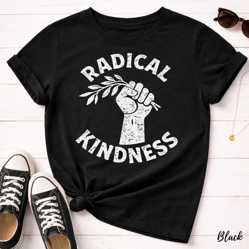May include: Black t-shirt with the words "RADICAL KINDNESS" in a white, distressed font. A white fist holds a sprig of leaves. The shirt is paired with black sneakers.