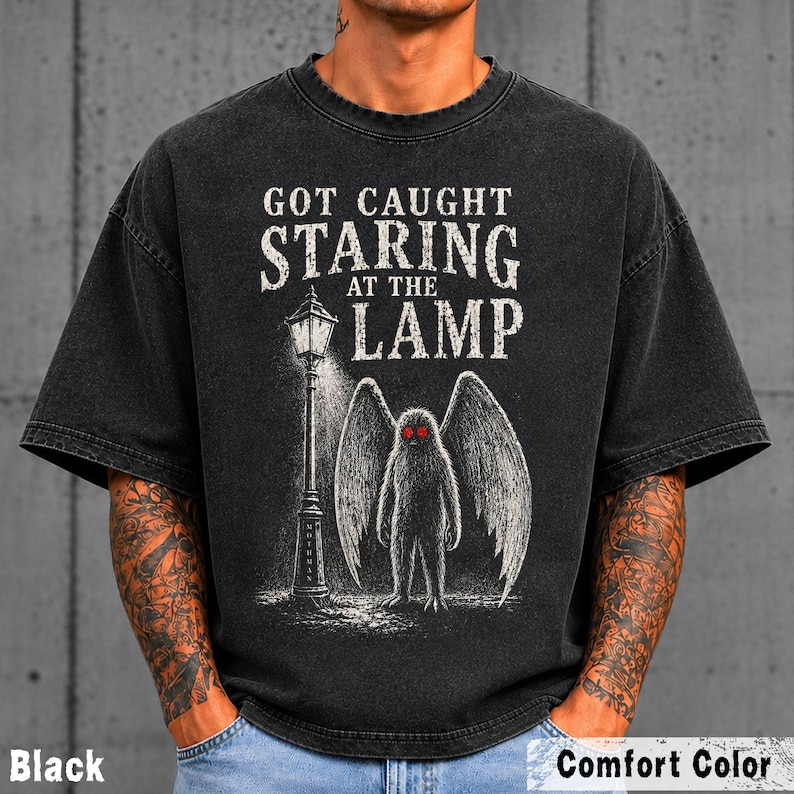 May include: A black short-sleeve t-shirt featuring a graphic print of a winged creature standing next to a street lamp. The text reads GOT CAUGHT STARING AT THE LAMP. The creature has bright red eyes and large wings, rendered in a white sketch style.