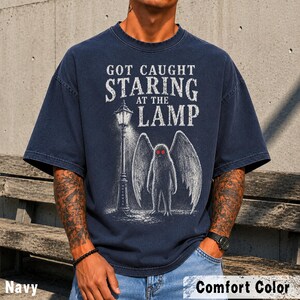 May include: A navy blue short-sleeve t-shirt featuring a white graphic print of a winged creature standing next to a street lamp. The text reads GOT CAUGHT STARING AT THE LAMP. The creature has two bright red eyes and large feathered wings.