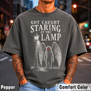 May include: A charcoal grey t-shirt featuring a white graphic of a winged creature standing next to a street lamp. The text reads GOT CAUGHT STARING AT THE LAMP. The design includes a small Mothman label on the lamp post and two bright red eyes.