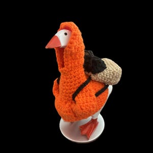 Könnte beinhalten: Dekorative Gänsefigur mit orangefarbener Häkelhaube und schwarzen Akzenten. Die Gans hat einen weißen Kopf, einen orangefarbenen Schnabel und rote Füße. Ein kleiner Rucksack ist am Rücken der Gans befestigt. Die Figur steht auf einem weißen Sockel.
