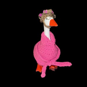 May include: A whimsical goose figurine wearing a pink robe, glasses, and a hat. The goose has an orange beak and feet. The hat is made of twine and adorned with pink flowers. The background is black.