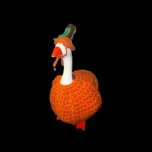May include: A white goose figurine wearing a textured orange crocheted sweater and a matching orange crocheted hat topped with a small green leaf accent. The goose has a bright orange beak and orange feet, standing against a solid black background.
