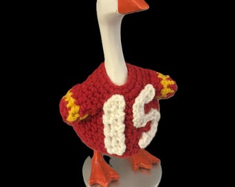 May include: A goose ornament with a white head, orange beak and feet. It is wearing a red crocheted sweater with the number 15 in white and yellow sleeve accents. The figurine stands on a silver base. A quirky decorative item.