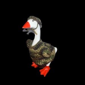 May include: A white goose figurine with a bright orange beak and orange webbed feet. The goose is wearing a small, hand-knitted sweater and a matching beret, both featuring a camouflage pattern in shades of dark green, brown, and black.