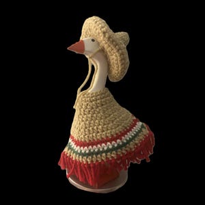 Peut inclure: Une figurine d'oie portant un sombrero et un poncho crochetés. Le sombrero beige a un large bord et une mentonnière. Le poncho est beige avec des rayures rouges, vertes et blanches et des franges rouges. L'oie a un corps blanc, un bec orange et est sur fond noir.
