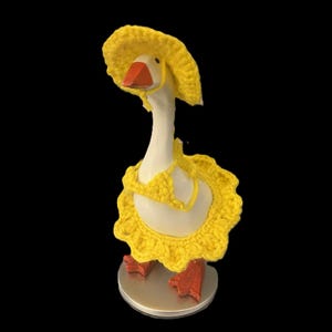 May include: A whimsical goose figurine wearing a yellow crocheted sun hat and matching ruffled collar. The goose has an orange beak and feet, standing on a silver base. The outfit adds a touch of humor and charm to the decorative piece.