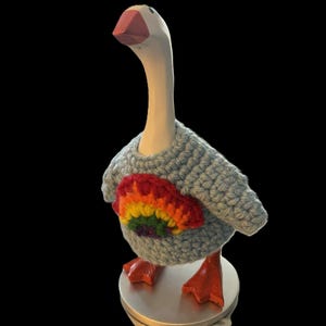 Può includere: Statuetta di oca in legno con un maglione all'uncinetto azzurro con un cuore arcobaleno. L'oca ha un collo bianco, un becco rosso e zampe arancioni. La statuetta è su una base argentata.