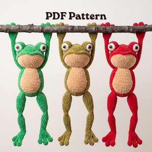 May include: Three crocheted frog plushies in green, olive, and red, each with a tan belly and large eyes, hanging from a horizontal wooden branch. The text PDF Pattern is displayed above the frogs. The plushies feature textured yarn.