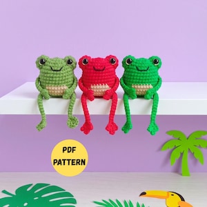 May include: Three crocheted frog dolls in light green, red, and dark green, each featuring large black eyes, a stitched smile, and a tan belly. The frogs have long dangling legs and arms. A yellow circular badge displays the text PDF PATTERN.