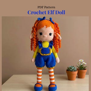 May include: A crochet elf doll with bright orange curly hair, blue bows, and pointed ears. The doll wears a yellow long-sleeved shirt, blue overalls with ruffled straps, and orange and white striped leggings with blue shoes. Text reads PDF Pattern Crochet Elf Doll.