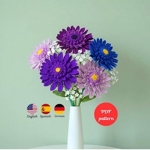 Puede incluir: Un ramo de flores de ganchillo hechas a mano en un jarrón blanco. Las flores son en tonos de morado, azul y lavanda con centros amarillos. El ramo incluye pequeñas flores blancas. Un círculo rojo con el texto "PDF pattern" es visible.